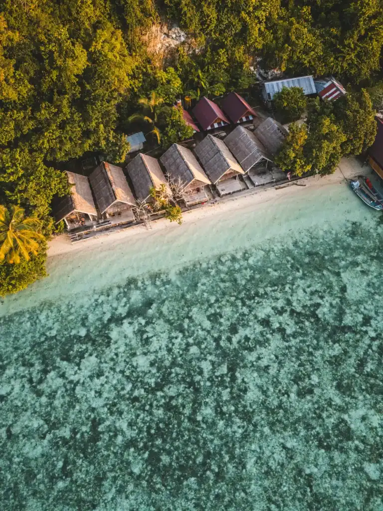 Aerial view of overwater homestay bungalows on Kri Island, Raja Ampat surrounded by turquoise reef and jungle