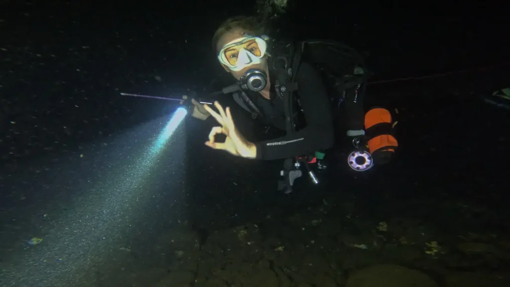 Diver giving OK signal during a night dive on the USAT Liberty Wreck in Tulamben, Bali, with torch beam illuminating volcanic sand.